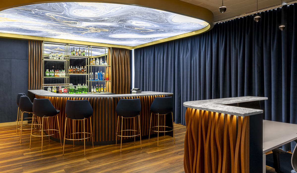 Bar interior de diseño elegante y contemporáneo. La barra principal tiene una forma curva y está revestida con paneles verticales de madera en tonos cálidos, iluminados desde la base con luz indirecta que resalta las ondulaciones del material. La encimera es de piedra gris con acabado pulido. Frente a la barra se alinean varios taburetes altos con estructura metálica dorada y asientos tapizados en azul oscuro. Detrás de la barra, una estantería iluminada exhibe diversas botellas de bebidas organizadas en estantes de vidrio, con un fondo espejado que amplía visualmente el espacio. El techo destaca por un panel ovalado con un diseño marmoleado en tonos blanco, gris y azul, enmarcado con una moldura dorada que integra iluminación perimetral. A la derecha, cortinas gruesas en azul marino cubren toda la pared, aportando textura y profundidad. El suelo es de madera en tonos cálidos, armonizando con el revestimiento de la barra. A un lado se aprecia una mesa alta auxiliar con el mismo acabado ondulado e iluminación integrada.