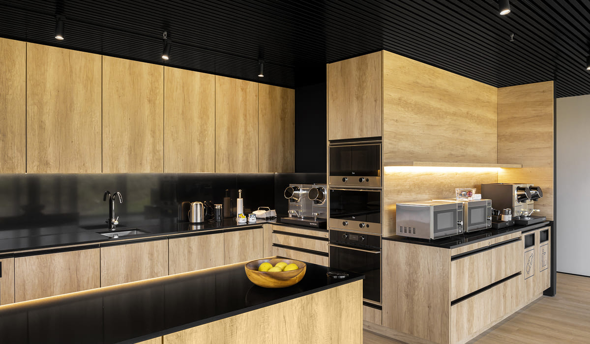 Cocina de oficina moderna con un diseño elegante y minimalista. Los muebles están revestidos en madera clara con vetas visibles, combinados con encimeras y salpicaderos en acabado negro brillante que aportan contraste. A la izquierda se observa un fregadero empotrado con grifería negra, junto a pequeños electrodomésticos como un hervidor eléctrico y una cafetera. En la parte central hay una columna con horno y microondas integrados en acero inoxidable. A la derecha, sobre otra encimera, se alinean dos microondas y una máquina de café adicional. Bajo esta zona se distinguen compartimentos señalizados para reciclaje. La iluminación LED indirecta bajo los armarios superiores y en la parte inferior de los muebles crea un ambiente cálido y sofisticado. En primer plano, una isla o barra baja sostiene un cuenco de madera con manzanas verdes. El techo es negro con iluminación puntual empotrada, reforzando el estilo contemporáneo del espacio.