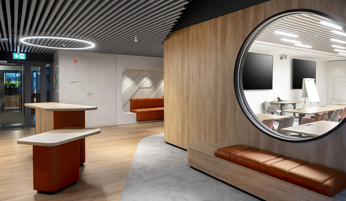 Espacio interior moderno con un diseño elegante y contemporáneo. A la derecha se aprecia una estructura curva revestida en madera clara con un gran ventanal circular tipo “ojo de buey” que permite ver una sala de reuniones equipada con mesas, sillas, una pizarra móvil y pantallas en la pared. Frente a esta estructura hay un banco integrado con cojines tapizados en tono marrón cuero. En el centro-izquierda del espacio se observan dos mesas altas de diseño orgánico con bases en color naranja y superficies en madera clara. Al fondo, hay una zona de espera con un sofá tapizado en el mismo tono marrón, empotrado en un nicho de pared con iluminación suave. El techo presenta lamas lineales en color oscuro y una luminaria circular suspendida que aporta un efecto moderno y sofisticado. El suelo combina madera clara con una zona alfombrada en gris, creando contraste y delimitando áreas dentro del espacio.