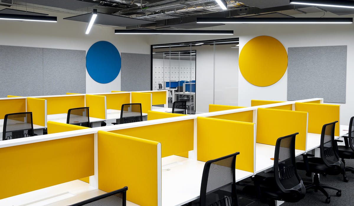 Oficina moderna con un diseño abierto tipo “open space”. Los puestos de trabajo están organizados en filas con escritorios blancos y paneles divisorios en color amarillo brillante, que aportan energía y dinamismo al ambiente. Cada estación cuenta con una silla ergonómica negra con respaldo de malla. En la pared del fondo se observan paneles acústicos grises y dos elementos decorativos circulares grandes, uno azul y otro amarillo, que refuerzan la identidad visual del espacio. Al centro hay una puerta corrediza de vidrio que conduce a otra sala con casilleros blancos y áreas adicionales de trabajo. El techo presenta un estilo industrial con conductos visibles, iluminación lineal suspendida en negro y luminarias en ángulo, lo que aporta un aire contemporáneo y tecnológico. En conjunto, el espacio transmite modernidad, organización y un entorno colaborativo.