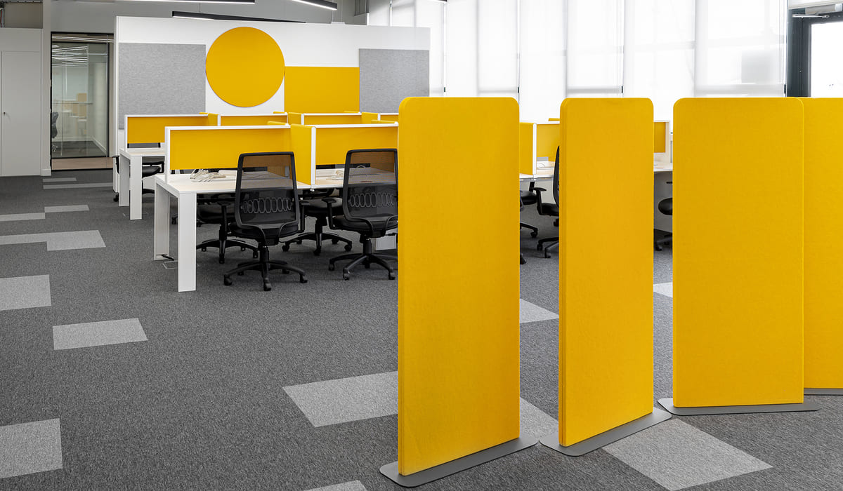 Espacio de oficina abierta con un diseño moderno y funcional. Se observan varios puestos de trabajo organizados en filas, con escritorios blancos y sillas ergonómicas negras con respaldo de malla. Cada estación está delimitada por paneles divisores en color amarillo vibrante, que aportan dinamismo y ayudan a crear separación visual y acústica entre los empleados. En primer plano destacan tres paneles acústicos independientes, también en amarillo, con base metálica gris, colocados como elementos de separación adicional. El suelo está cubierto con moqueta gris en distintos tonos formando un patrón geométrico sutil. Al fondo, la pared combina paneles grises con un gran círculo decorativo amarillo que refuerza la identidad cromática del espacio. La iluminación es uniforme y natural, complementada con luminarias lineales en el techo. El conjunto transmite una estética contemporánea, colaborativa y enérgica, ideal para entornos de trabajo dinámicos.