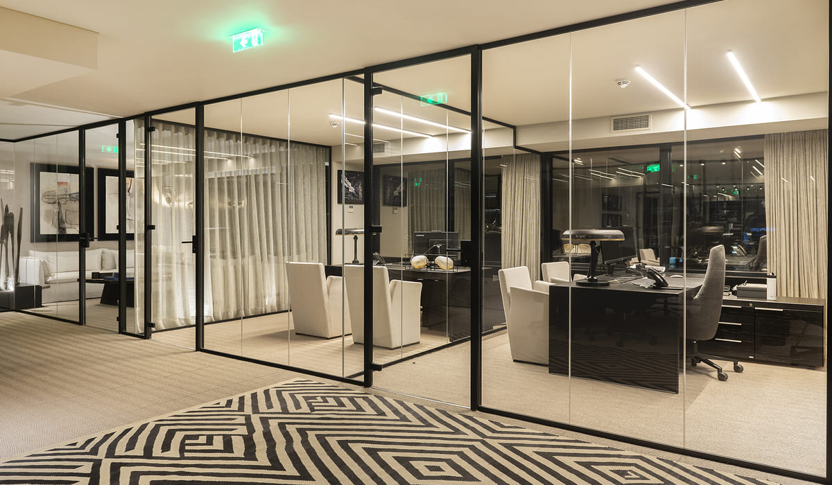Oficina ejecutiva de estilo moderno, delimitada por mamparas de vidrio con marcos negros. En el interior se observa un escritorio amplio en acabado oscuro con una lámpara de diseño, teléfono y elementos decorativos. Frente al escritorio hay varias sillas tapizadas en color claro, mientras que detrás se encuentra una silla ejecutiva de respaldo alto en tono gris. El espacio está iluminado con luz cálida y líneas de iluminación empotradas en el techo, lo que genera un ambiente elegante y profesional. Las cortinas claras cubren parte de los ventanales, suavizando la entrada de luz exterior. En el suelo se aprecia una moqueta beige y, en primer plano, una alfombra con patrón geométrico en blanco y negro. Al fondo, se distinguen otras salas acristaladas y un área tipo lounge con sofá y decoración artística en las paredes, reforzando la estética contemporánea y sofisticada del entorno.
