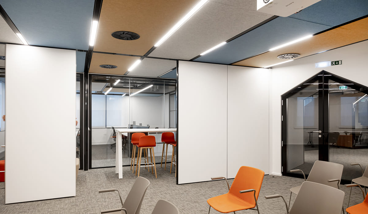 Espacio de oficina moderno con diseño contemporáneo. En primer plano se ven varias sillas de espera en tonos neutros y naranja, dispuestas sobre una alfombra gris. A la derecha hay una pequeña sala cerrada con estructura de vidrio y marco negro, con puerta también acristalada, que permite ver el interior. En el centro se observa otra sala delimitada por paneles blancos y vidrio. Dentro hay una mesa alta blanca acompañada de taburetes altos de color naranja con patas de madera, lo que aporta un contraste cálido al entorno. El techo combina paneles acústicos en tonos beige y azul grisáceo, con iluminación lineal empotrada y rejillas de ventilación circulares. El conjunto transmite un ambiente profesional, funcional y bien iluminado, pensado para reuniones informales y trabajo colaborativo.