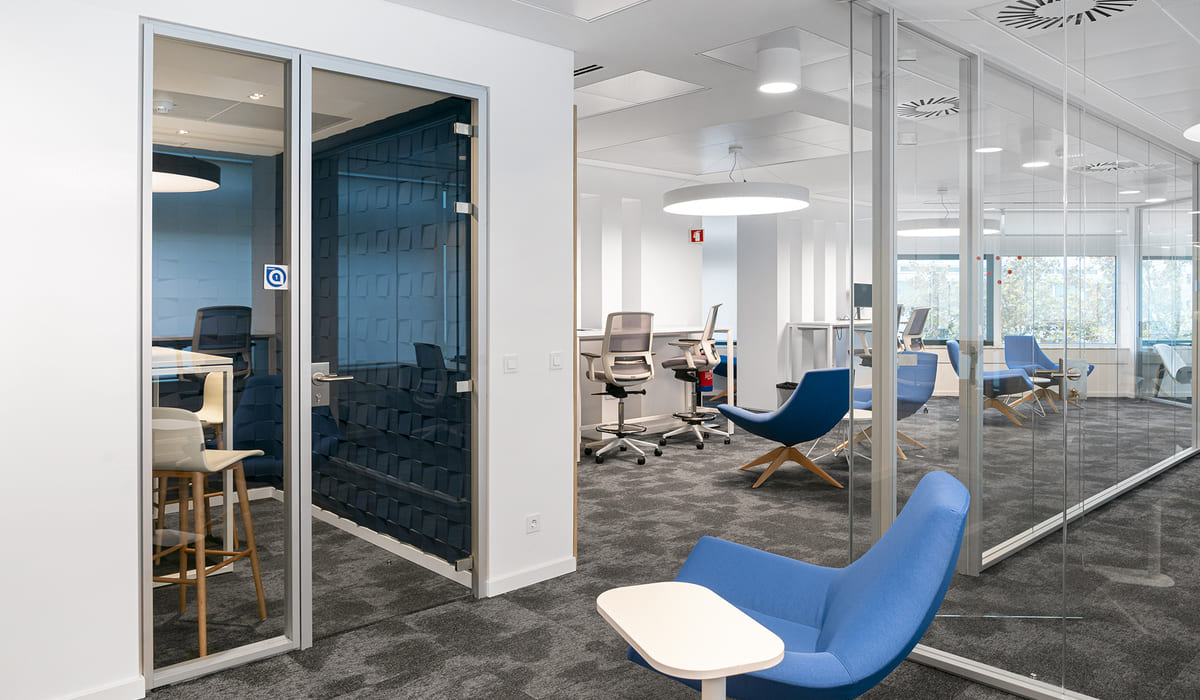 Espacio de oficina contemporáneo con divisiones acristaladas que delimitan varias salas y áreas de trabajo. En primer plano, a la derecha, se observa una silla lounge tapizada en azul con una pequeña mesa auxiliar blanca integrada en el lateral. El suelo está cubierto con moqueta gris de textura uniforme. A la izquierda se ve una pequeña sala cerrada con paredes de vidrio y una puerta con marco metálico. En su interior hay una mesa alta con taburetes de madera y asiento blanco. Junto a esta sala, una pared decorativa en tono azul oscuro presenta un relieve geométrico tridimensional que aporta profundidad visual. Al fondo, el espacio se abre hacia una zona de trabajo compartida con escritorios blancos y sillas ergonómicas en tonos neutros. También se distinguen más sillones azules distribuidos estratégicamente para reuniones informales. El techo blanco incorpora luminarias circulares empotradas y sistemas de ventilación visibles, mientras que grandes ventanales permiten la entrada de luz natural.