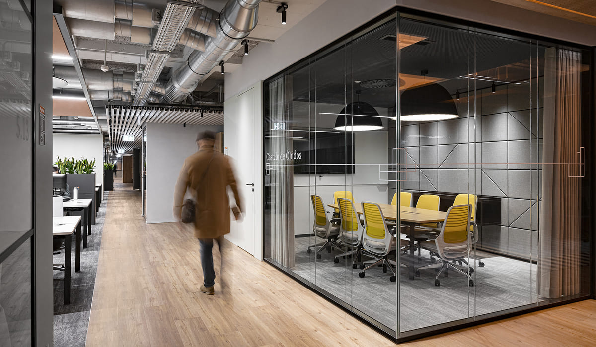 Pasillo de oficina moderna con un diseño industrial y contemporáneo. A la derecha se observa una sala de reuniones cerrada con paredes de vidrio y marcos negros. En su interior hay una mesa rectangular de madera rodeada por varias sillas ergonómicas con respaldo amarillo y estructura blanca. La pared del fondo está revestida con paneles acústicos grises con diseño geométrico, y una lámpara colgante negra ilumina la mesa. En el pasillo, el suelo es de madera clara y el techo deja a la vista conductos metálicos y sistemas de ventilación, reforzando el estilo industrial. A la izquierda se distinguen escritorios de trabajo con ordenadores y plantas decorativas. Una persona aparece caminando por el pasillo, ligeramente desenfocada, lo que aporta sensación de movimiento y actividad en el entorno laboral.