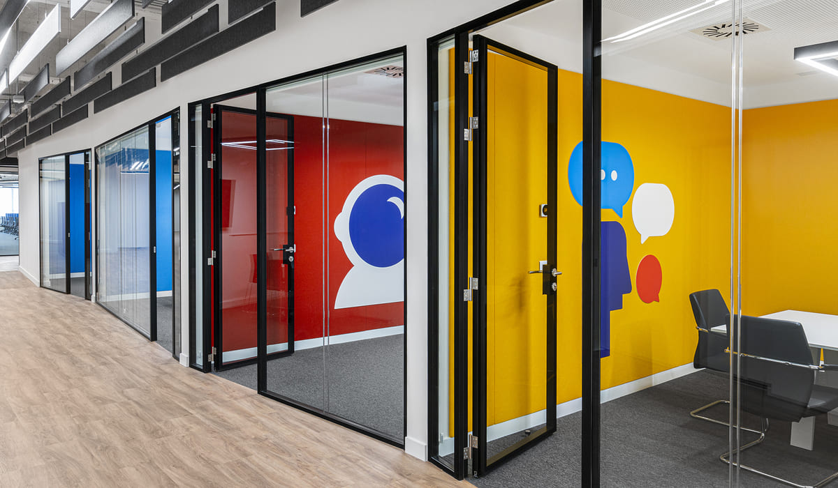Pasillo de oficina moderno con varias salas de reuniones cerradas por paredes y puertas de vidrio con marcos negros. Cada sala tiene una pared interior de color llamativo: una en rojo con un ícono blanco y azul de perfil humano, y otra en amarillo con ilustraciones de siluetas y globos de diálogo en azul y rojo, lo que sugiere espacios destinados a la comunicación y colaboración. El suelo es de madera clara en el pasillo y moqueta gris dentro de las salas. En el interior de las salas se observan mesas blancas y sillas negras de diseño sencillo. El techo presenta paneles acústicos y luminarias lineales, reforzando el estilo contemporáneo y funcional del espacio.