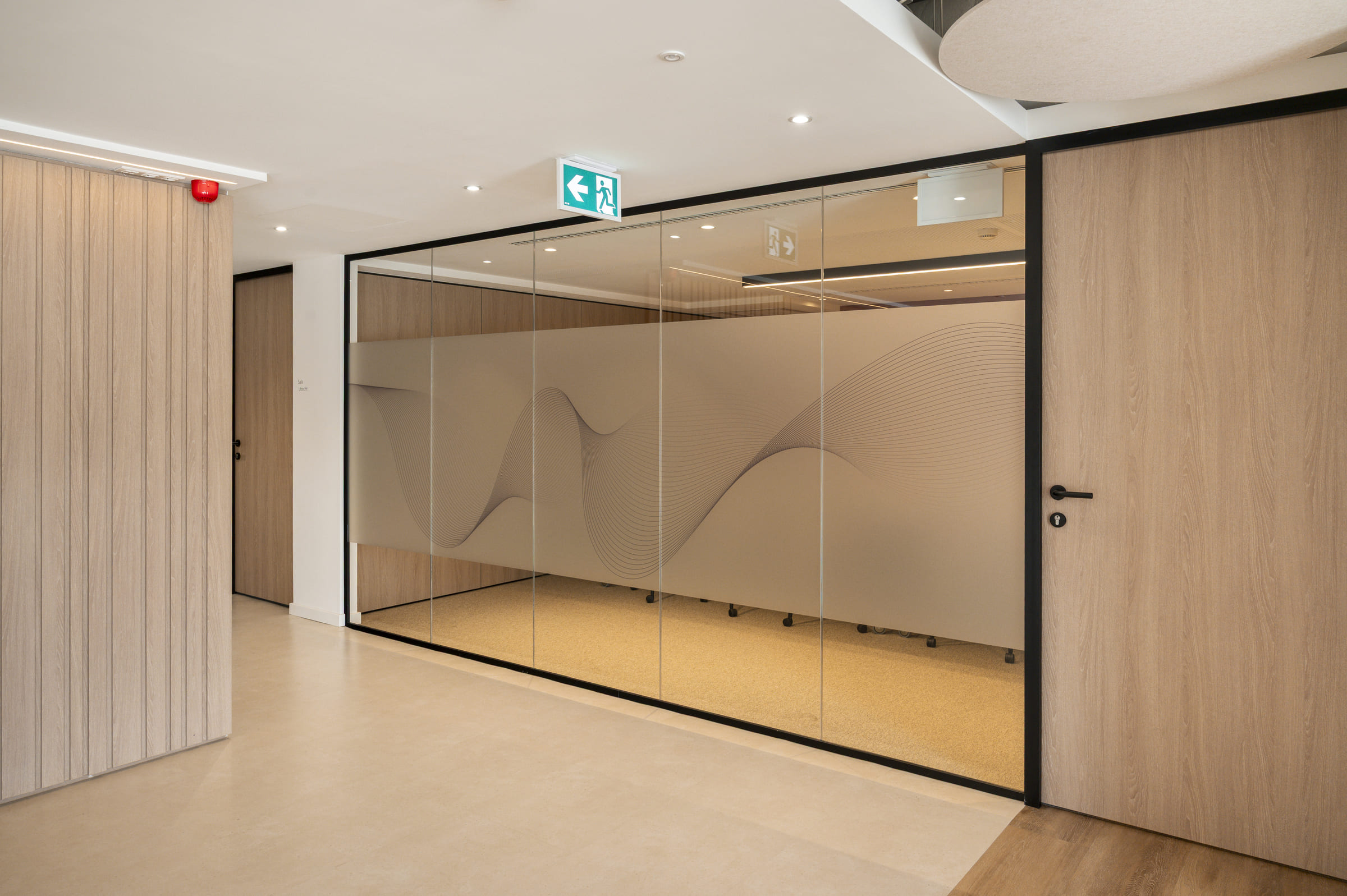 Pasillo de oficina con un diseño moderno y minimalista. A la derecha se observa una sala delimitada por una mampara de vidrio con marco negro. La parte central del vidrio está cubierta con una lámina translúcida en tono beige que incorpora un diseño gráfico de líneas onduladas, generando privacidad parcial sin bloquear completamente la luz. Dentro de la sala se distinguen las bases con ruedas de varias sillas alrededor de una mesa, sobre un suelo alfombrado en color claro. En el techo hay iluminación empotrada y una señal verde de salida de emergencia con flecha hacia la izquierda. Las paredes y puertas del pasillo están revestidas en madera clara, con manillas negras que contrastan con el acabado natural. El suelo del pasillo es liso y beige, mientras que el interior de la sala tiene un revestimiento diferente, creando una separación visual entre ambos espacios. El conjunto transmite orden, sobriedad y un estilo corporativo contemporáneo.