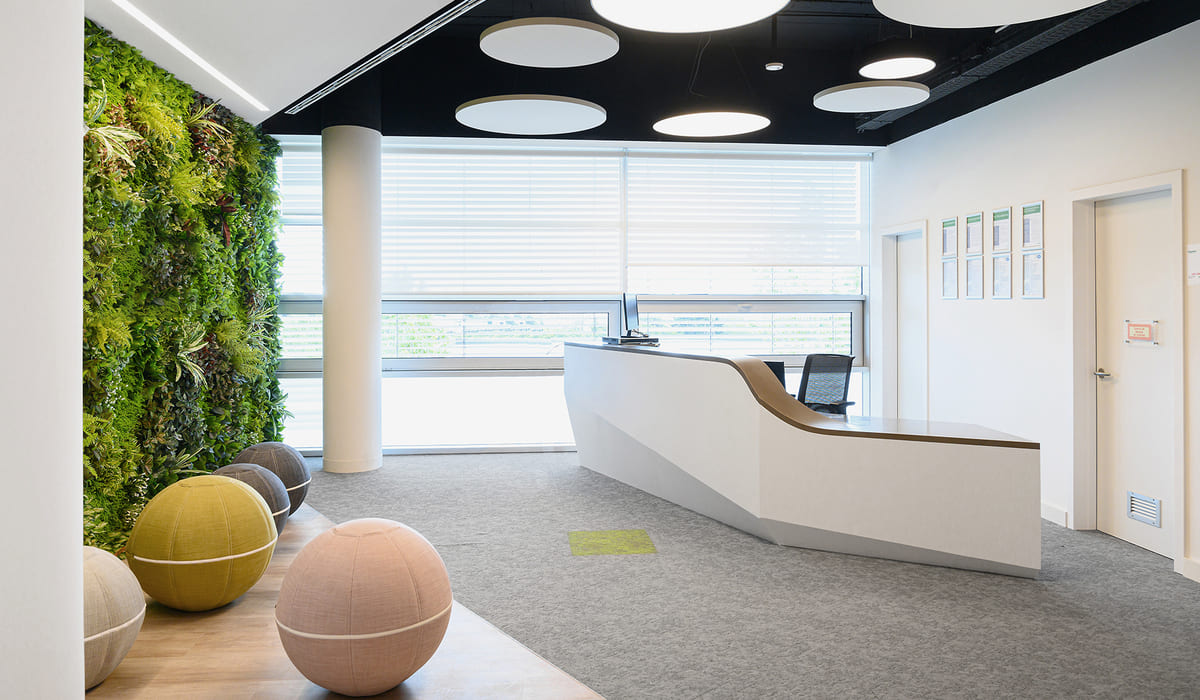 Recepción moderna y luminosa con un diseño contemporáneo. A la derecha se encuentra un mostrador blanco de líneas curvas y minimalistas, con una superficie superior en tono madera clara. Detrás del mostrador hay una silla de oficina y, en la pared lateral, varios marcos alineados que parecen contener certificados o documentos. A la izquierda destaca un jardín vertical cubierto de vegetación natural que aporta frescura y un contraste vibrante con las superficies blancas. Frente a este muro verde hay varios asientos esféricos tapizados en tonos neutros como beige, mostaza y gris. El techo es negro y cuenta con luminarias circulares blancas empotradas que distribuyen la luz de manera uniforme. Al fondo, grandes ventanales con persianas translúcidas permiten la entrada de luz natural, reforzando la sensación de amplitud y limpieza del espacio.