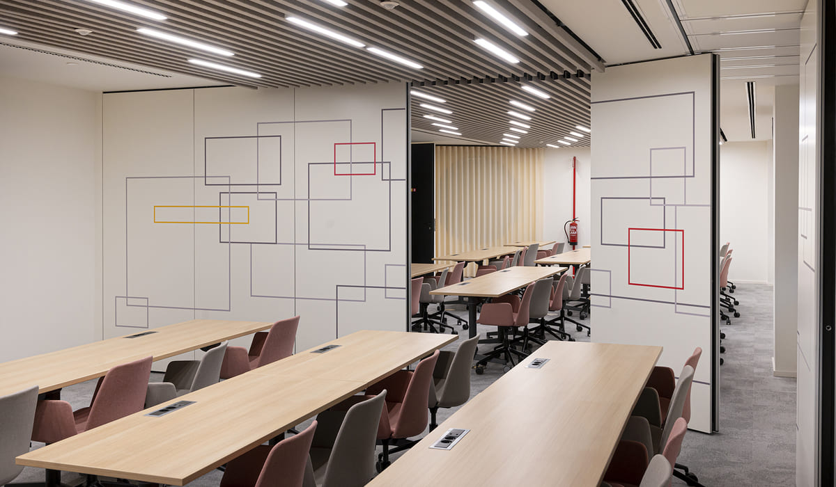 Sala de formación o espacio de trabajo colaborativo dentro de una oficina moderna. El área cuenta con varias mesas rectangulares de madera clara dispuestas en filas paralelas, acompañadas de sillas tapizadas en tonos suaves como rosa y gris, todas con ruedas para facilitar la movilidad. Las paredes blancas están decoradas con un diseño gráfico minimalista compuesto por líneas finas que forman rectángulos superpuestos en distintos tamaños, algunos resaltados en colores como rojo y amarillo, aportando un toque dinámico y contemporáneo al espacio. El techo presenta un diseño lineal con lamas y luminarias LED integradas que ofrecen iluminación uniforme. Al fondo se aprecia una división móvil o panel deslizante que conecta con otra sala similar, ampliando la capacidad del espacio. También se distingue un extintor de incendios en la pared posterior. El suelo es de moqueta en tono gris, lo que aporta confort acústico y visual.
