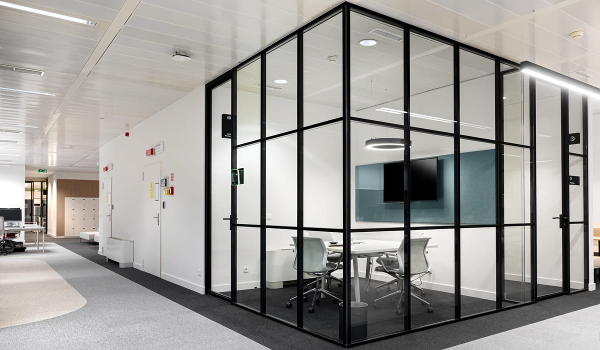 Oficina moderna con una sala de reuniones completamente cerrada por paredes de vidrio con perfilería metálica negra. En el interior se aprecia una mesa rectangular blanca rodeada de sillas ergonómicas grises con ruedas, así como una pantalla plana montada en la pared frontal. La iluminación es uniforme y contemporánea, con luminarias circulares y lineales en el techo. El pasillo exterior combina zonas de moqueta gris con superficies claras, y al fondo se observan puestos de trabajo abiertos y armarios, creando un entorno profesional, ordenado y funcional.
