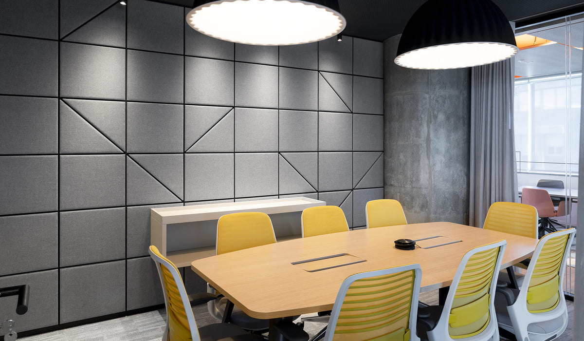 Sala de reuniones moderna con un diseño elegante y sobrio. La pared principal está revestida con paneles acústicos grises dispuestos en un patrón geométrico, lo que aporta textura y mejora la acústica del espacio. En el centro se sitúa una mesa rectangular de madera clara con canaletas integradas para cableado, rodeada de sillas ergonómicas con estructura blanca y asientos amarillos, que aportan un toque de color. La iluminación se compone de dos lámparas colgantes de diseño contemporáneo, una blanca y otra negra, que proporcionan una luz cálida y uniforme. El conjunto transmite un ambiente profesional, cómodo y bien cuidado, ideal para reuniones de trabajo.