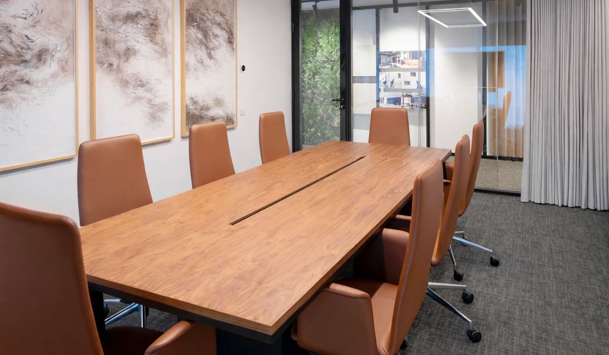 Sala de reuniones moderna con una mesa rectangular de madera en tono cálido y acabado natural. La mesa incorpora una ranura central para gestión de cableado, lo que indica funcionalidad tecnológica para reuniones formales o presentaciones. Alrededor de la mesa se disponen varias sillas tapizadas en cuero o material similar en color marrón caramelo, con respaldo alto y base metálica con ruedas, aportando comodidad y movilidad. En la pared izquierda se observan tres cuadros de gran formato con arte abstracto en tonos neutros, enmarcados en madera clara, que aportan elegancia al espacio. Al fondo, una puerta de vidrio con marco negro conecta con otra área de la oficina, y junto a ella hay cortinas claras que suavizan la iluminación. También se aprecia una pared vegetal en la zona contigua, añadiendo un elemento natural al entorno corporativo. El conjunto transmite una atmósfera profesional, sofisticada y acogedora, ideal para reuniones ejecutivas.