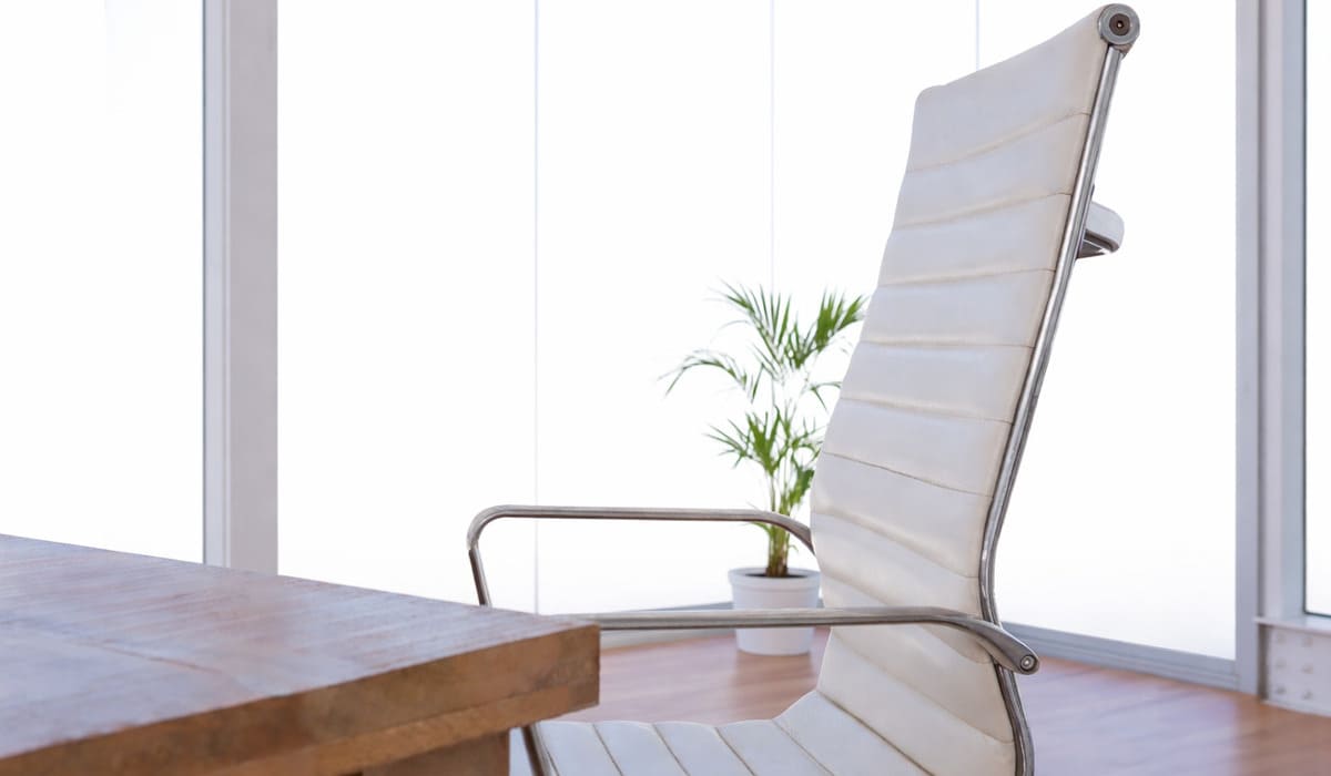Silla de oficina ejecutiva tapizada en color blanco, con respaldo alto y diseño ergonómico. El respaldo presenta costuras horizontales acolchadas que le aportan un estilo elegante y contemporáneo. La estructura y los apoyabrazos son metálicos, con acabado cromado, lo que refuerza su estética moderna y profesional. En primer plano se observa parcialmente una mesa de madera maciza con acabado natural, de líneas rectas y diseño robusto. El entorno es luminoso, con grandes ventanales que permiten la entrada de luz natural. Al fondo, se aprecia una planta en maceta blanca que añade un toque decorativo y natural al espacio. El conjunto transmite una sensación de oficina moderna, limpia y minimalista, con una combinación equilibrada entre madera, metal y tonos neutros.