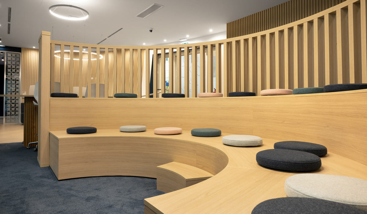 Espacio interior moderno con un diseño orgánico y acogedor. Destaca una estructura semicircular de madera clara que funciona como graderío o zona de asientos escalonados. Sobre los bancos de madera se distribuyen cojines redondos en tonos neutros —gris, beige, azul y verde suave— que aportan comodidad y un toque decorativo. En la parte superior, una celosía vertical de listones de madera sigue la curvatura del espacio, creando una separación visual ligera sin cerrar completamente el ambiente. La iluminación es cálida y uniforme, con luminarias empotradas en el techo y una lámpara circular suspendida que refuerza la geometría curva del diseño. El suelo combina madera y moqueta en tonos oscuros, delimitando áreas y aportando contraste. El conjunto transmite una sensación de calidez, diseño contemporáneo y funcionalidad, ideal para reuniones informales, presentaciones o espacios colaborativos.