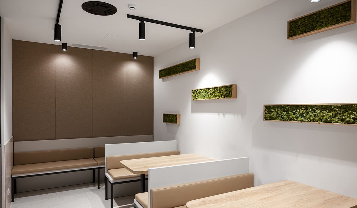 Pequeño espacio tipo comedor o zona de descanso dentro de una oficina moderna. Hay dos mesas rectangulares de madera clara con bancos tapizados en tono beige a ambos lados, creando un ambiente acogedor e informal. En la pared izquierda se observa un panel marrón que aporta calidez al espacio, mientras que la pared derecha está decorada con varias jardineras rectangulares de madera con vegetación verde, colocadas de forma horizontal a diferentes alturas, lo que añade un toque natural y decorativo. El techo es blanco y cuenta con iluminación mediante focos negros direccionables y un sistema de ventilación visible. El conjunto transmite un ambiente moderno, tranquilo y pensado para pausas, reuniones informales o comidas rápidas dentro de la oficina.