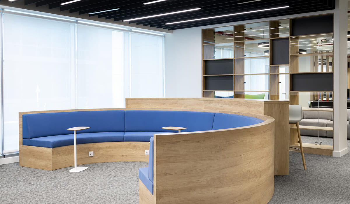 Espacio interior de oficina con diseño contemporáneo y enfoque colaborativo. En primer plano destaca un banco semicircular integrado, revestido en madera clara, con asiento acolchado en color azul. La estructura curva crea un ambiente más íntimo dentro de un espacio abierto. Frente al banco hay pequeñas mesas auxiliares redondas con base central blanca. El suelo está cubierto con moqueta gris, y al fondo se observa un módulo de estanterías abiertas en madera con compartimentos irregulares y algunos paneles oscuros, lo que aporta dinamismo visual. A través de los estantes se pueden ver otras áreas de la oficina con mobiliario moderno. A la izquierda, grandes ventanales con estores translúcidos permiten la entrada de luz natural difusa. El techo presenta lamas negras paralelas con iluminación lineal integrada, reforzando el estilo moderno y corporativo del espacio.