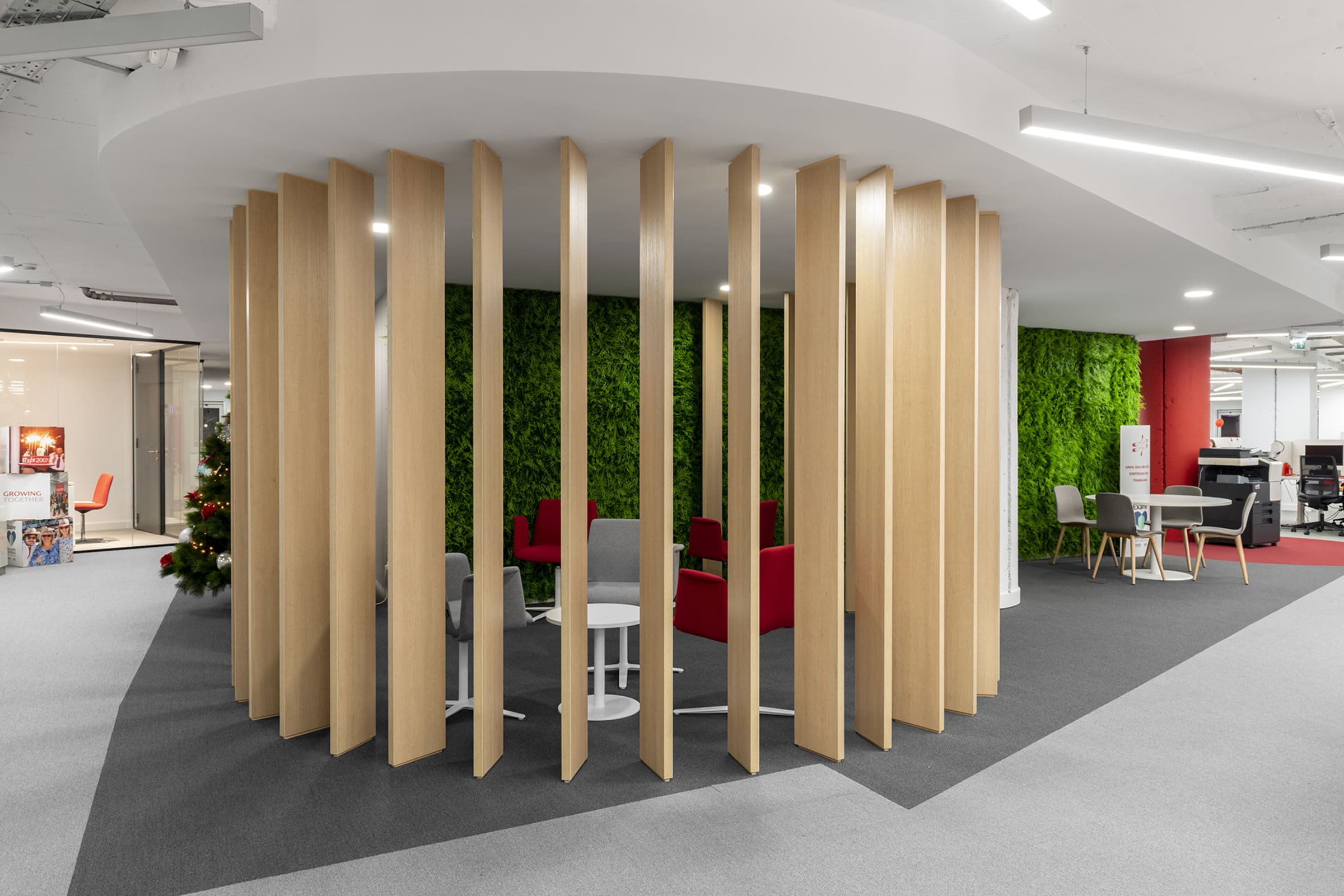 Espacio de oficina moderno concebido como una zona de descanso o reunión informal. El elemento central es una estructura circular formada por lamas verticales de madera clara, que delimita visualmente el área sin cerrarla por completo. En el interior se aprecian sillones tapizados en tonos grises y rojos, acompañados de mesas auxiliares blancas, creando un ambiente cómodo y colaborativo. El fondo está revestido con un muro vegetal verde, que aporta frescura y un contraste natural con los materiales neutros del entorno. El espacio es amplio, luminoso y de diseño contemporáneo, integrándose de forma armónica en una oficina abierta.