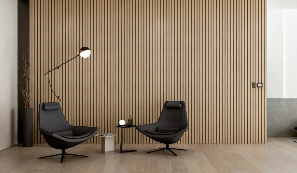 Espacio interior de diseño minimalista y elegante. Dos sillones modernos de color negro con base giratoria están dispuestos frente a una pared de listones verticales de madera clara. Entre ellos hay dos mesas pequeñas: una negra con una lámpara esférica encendida y otra más baja de color claro con una revista o libro. A la izquierda se aprecia una lámpara de pie con brazo articulado y pantalla redonda blanca, así como un jarrón alto y oscuro con ramas secas decorativas. El suelo es de madera en tonos naturales, creando un ambiente cálido y sofisticado.