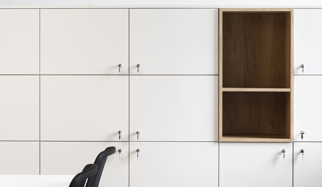 Pared con una estructura de casilleros blancos de diseño minimalista, cada uno con una pequeña cerradura y llave. En el centro derecho destaca un módulo abierto de madera con dos compartimentos, que rompe la uniformidad del conjunto y añade un toque cálido al diseño. En la parte inferior izquierda se observan parcialmente dos sillas negras junto a una mesa blanca, lo que sugiere un entorno de oficina o espacio de trabajo ordenado y funcional.