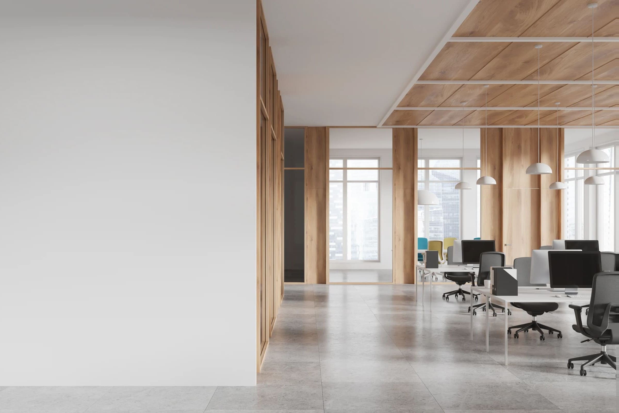 Oficina abierta y moderna con un diseño minimalista, donde predominan los tonos claros y la madera natural. A la izquierda, varias estaciones de trabajo con escritorios blancos y pantallas de ordenador negras están alineadas, cada una con su silla ergonómica gris. El techo presenta paneles de madera enmarcados por una estructura blanca y lámparas colgantes blancas de estilo contemporáneo. Grandes ventanales permiten la entrada abundante de luz natural y ofrecen una vista de edificios urbanos. Al fondo, se aprecian sillas de colores junto a otra zona de trabajo o reunión. A la derecha, una pared blanca lisa y paneles de madera dividen los espacios.