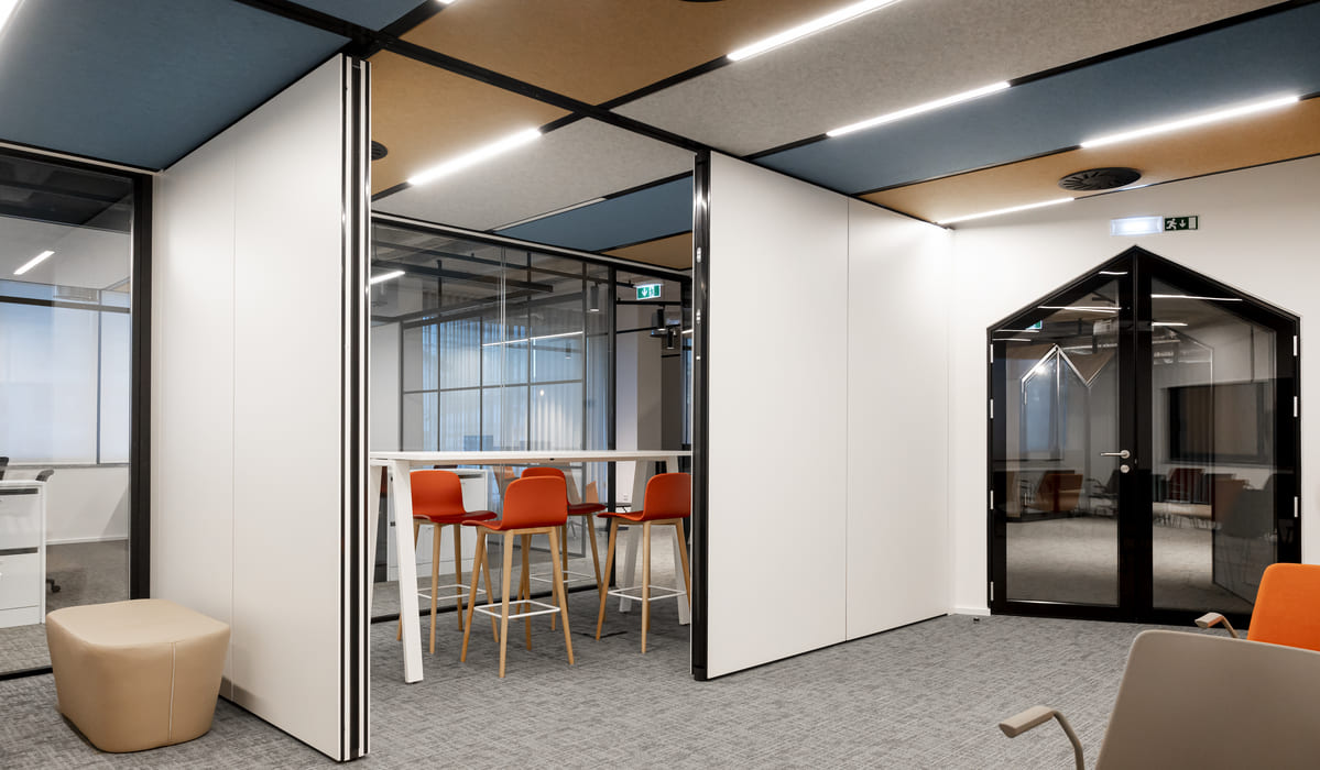 Espacio de oficina moderno y flexible, diseñado con un estilo contemporáneo. Se observan paneles móviles blancos que permiten dividir o abrir el espacio según las necesidades, combinados con mamparas de vidrio y marcos negros. En el centro hay una mesa alta con varias sillas altas de color naranja, pensada para reuniones informales o trabajo colaborativo. El techo presenta paneles acústicos en tonos beige y azul, integrados con líneas de iluminación LED que aportan una iluminación uniforme y elegante. El suelo está cubierto con moqueta gris, lo que refuerza la sensación de confort y aislamiento acústico. Al fondo destaca una puerta acristalada con forma geométrica, que aporta un toque distintivo al diseño general. El ambiente transmite funcionalidad, orden y modernidad.